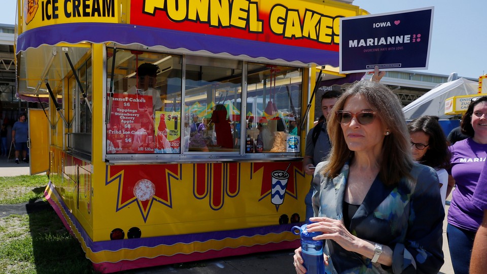 Marianne Williamson walks through the Iowa State Fair with a supporter holding a poster behind her as they walk in front of a funnel cake stand.