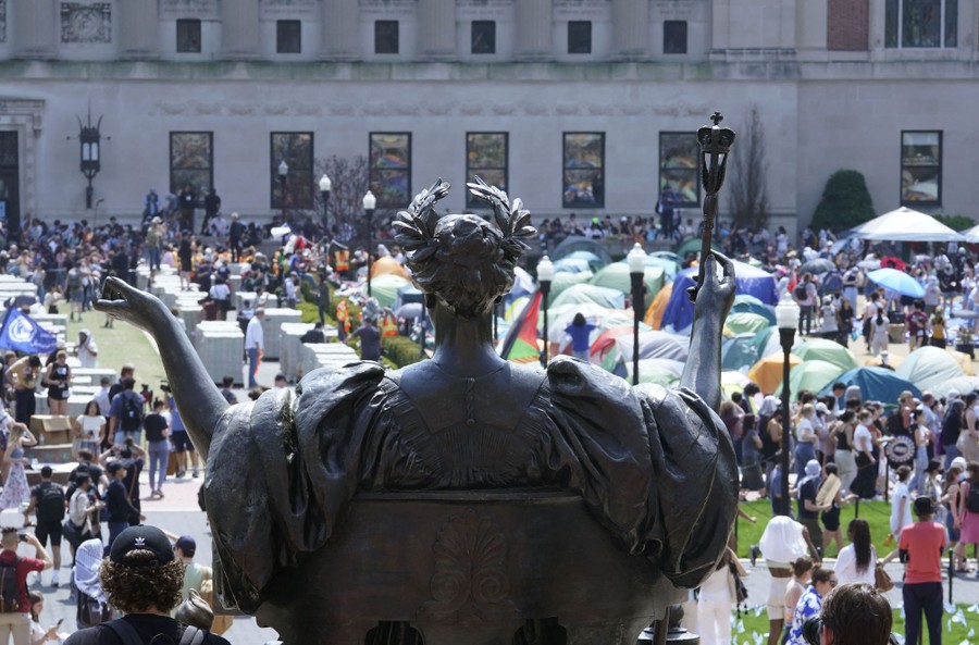 A view, looking past a statue, of many tents and people spread around a square on a college campus.