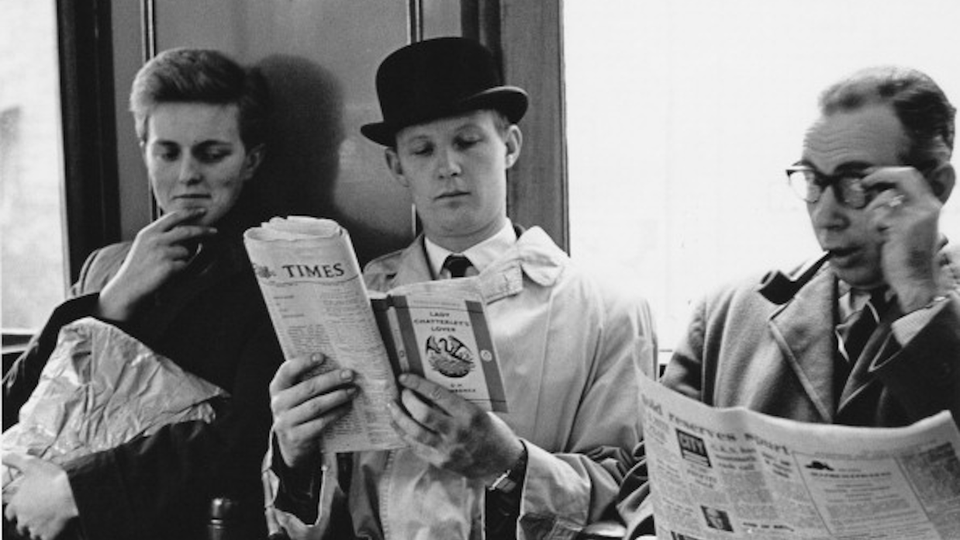 3 passengers on the train, one smirking over at another reading Lady Chatterley's Lover over the Times newspaper.