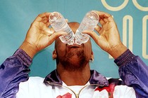 A man drinks from two plastic water bottles simultaneously. Both bottles are tipped over his mouth, blocking the upper half of his face.