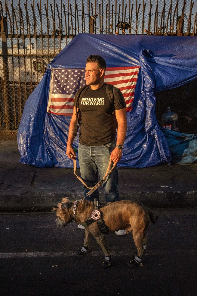 Man wearing "Moving Forward" t-shirt and jeans and holding leash of a dog wearing harness and booties in front of blue-tarp-covered tent with American flag