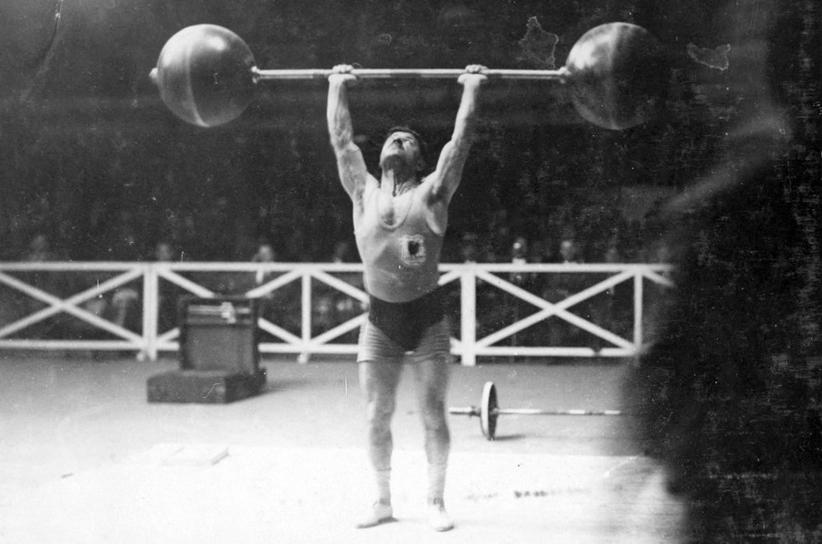 A weightlifter holds an old-fashioned barbell over his head.
