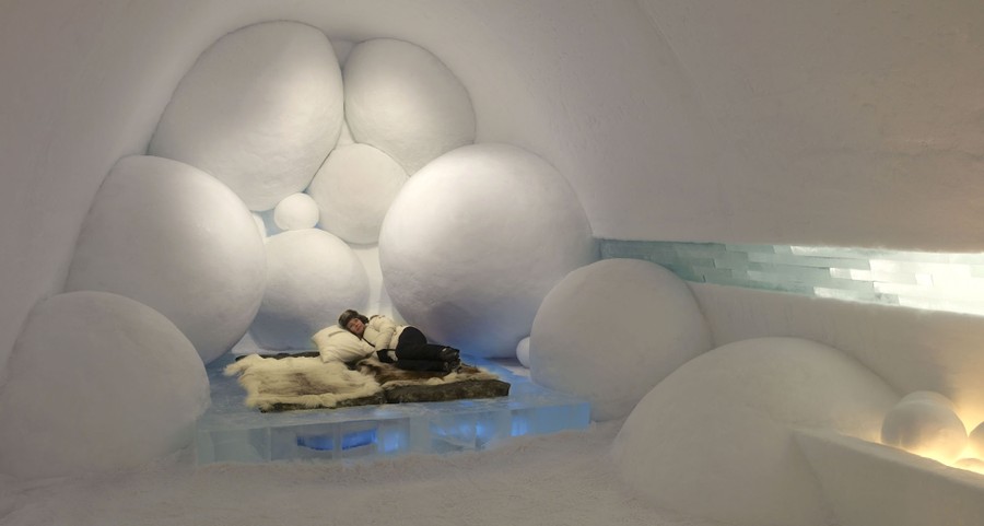 Một người nằm trên chiếc giường làm từ những khối băng phủ da tuần lộc, bên trong căn phòng chạm khắc bằng tuyết được trang trí bằng nhiều quả cầu chạm khắc lớn.