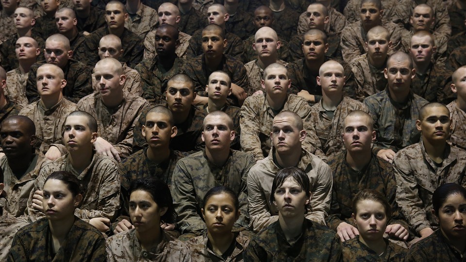 Marine recruits sit in a group listening to a speaker off camera.