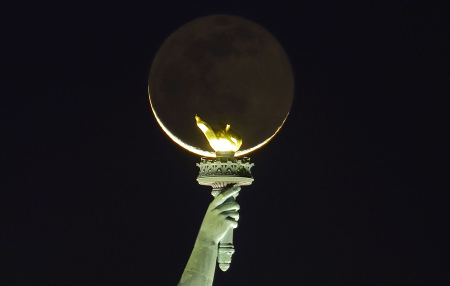 The moon rises behind the torch of the Statue of Liberty.