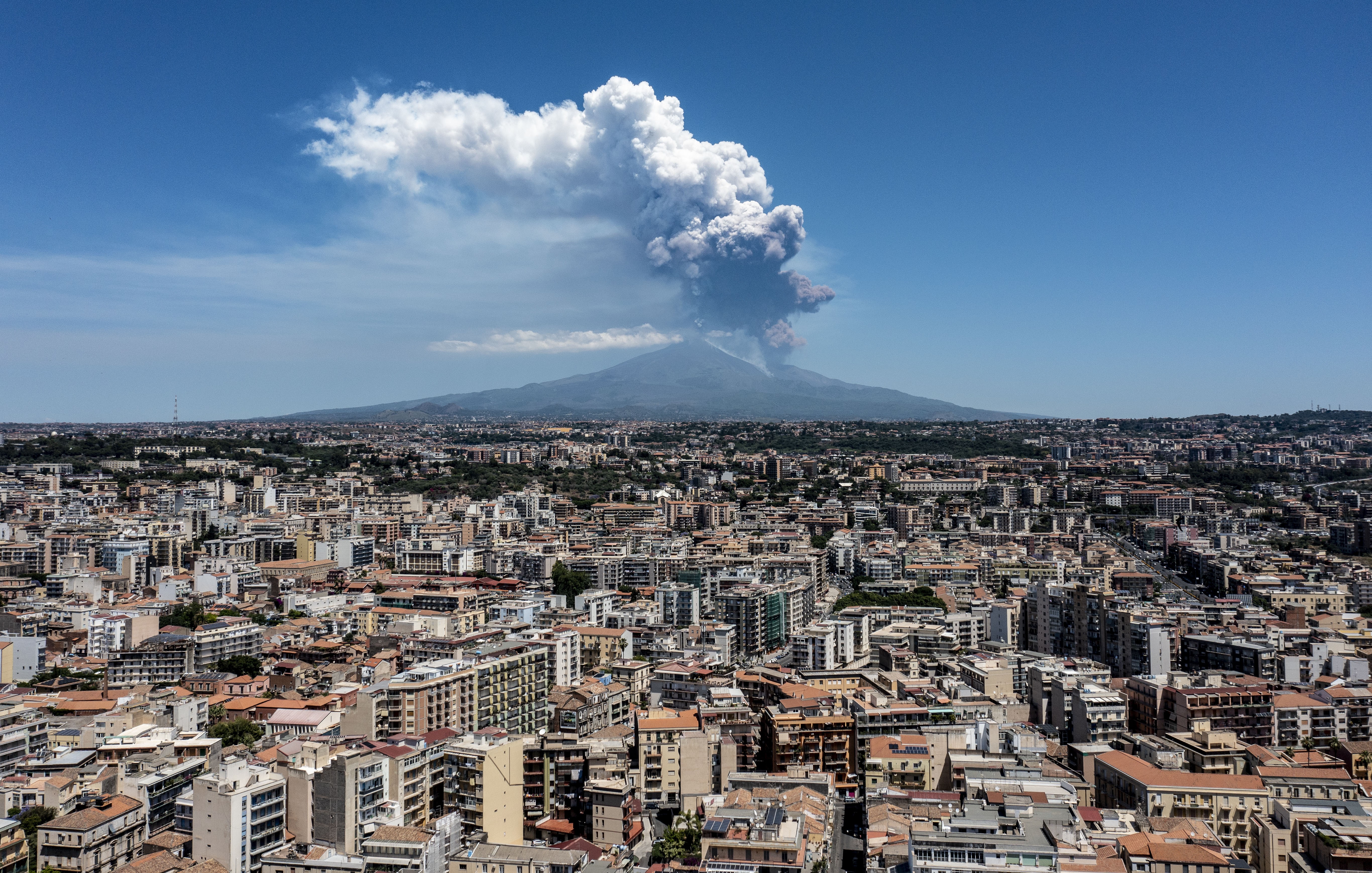 Ash and steam rise from the top of a volcano, with an aerial view of a city in the foreground.