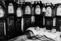 A man and woman sleep in a room full of antique clocks.