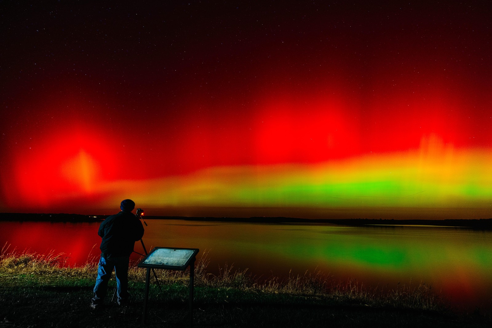 A photographer sets up a camera beside a lake at night, with a red and green aurora in the sky beyond.