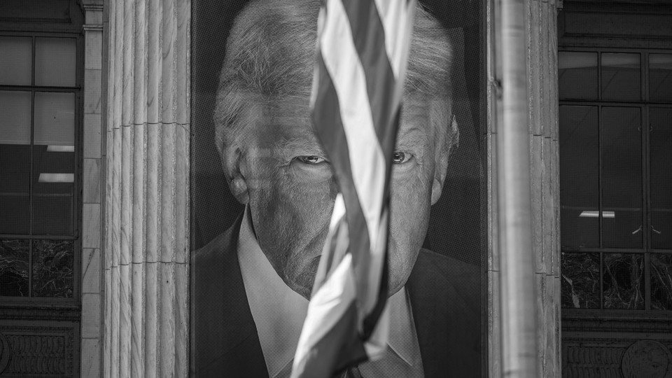 A banner with President Trump's image on it, hanging between building columns with an American flag flying between his eyes.