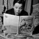 A black-and-white photo of a boy wearing a black shirt and peering at a newspaper comic, with newspapers stacked on both sides