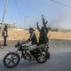 In a stretch of desert with smoke rising in the distance, a Hamas fighter on the back of a motorcycle raises his weapon and points to the sky.