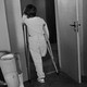 Black-and-white photo of a person standing with crutches in the doorway of a room containing prosthetic legs.