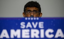 Dinesh D'Souza stands behind podium that reads: Save America. His glasses are spotted with rain.
