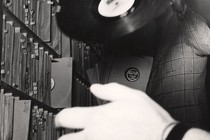 A black-and-white photo of a man in a plaid suit holding up a record, his face obscured