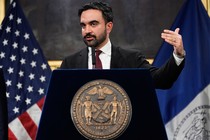 A photo of Mayor Zohran Mamdani speaking at a podium in front of an American flag and a New York City flag.