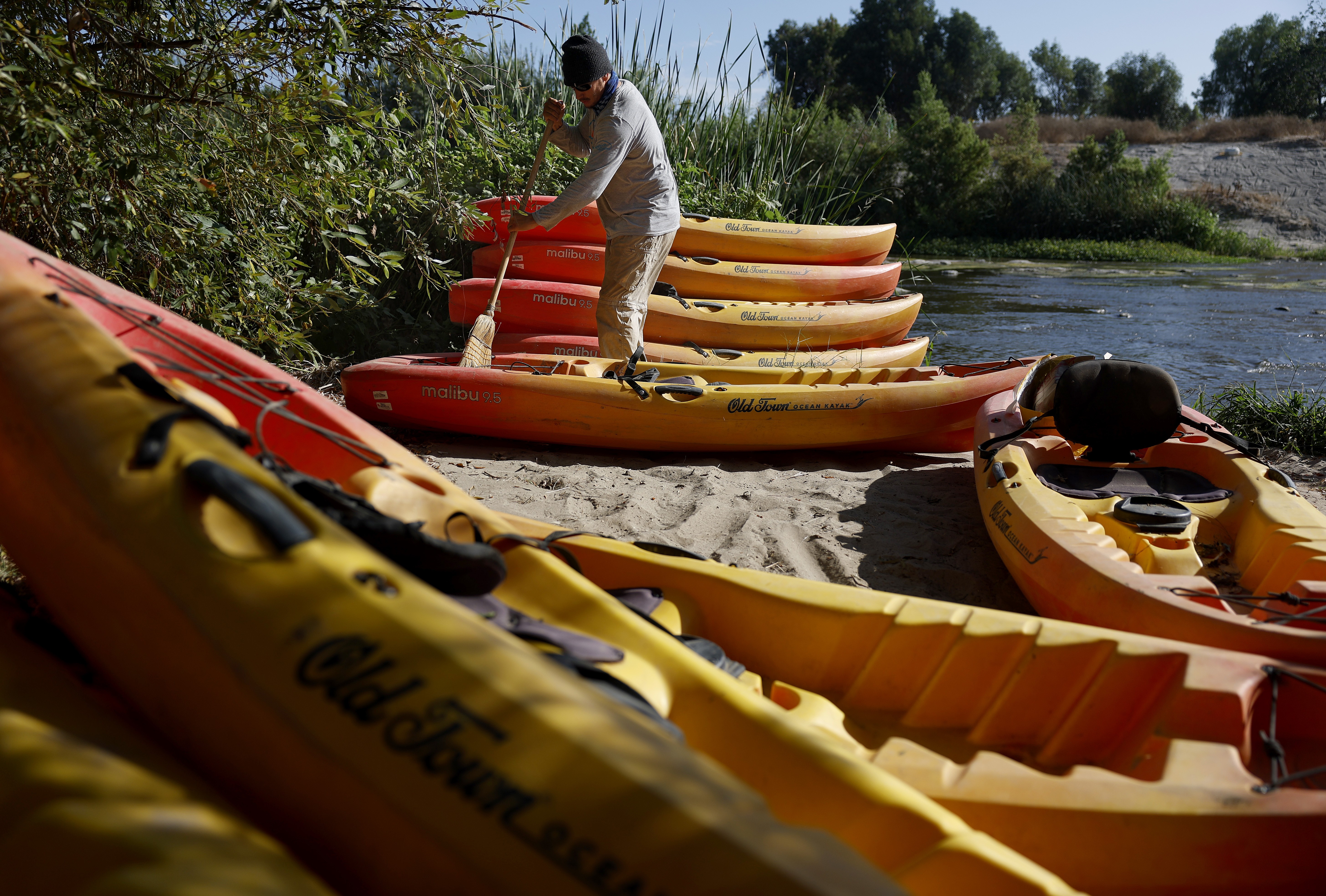 A person uses a broom to clean stacked kayaks beside a river.
