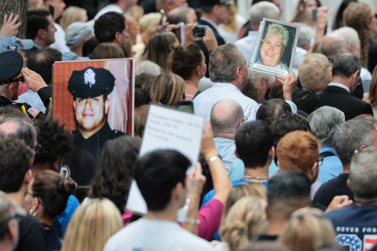A crowd of people, some holding signs with photos of loved ones, sit during a memorial ceremony.