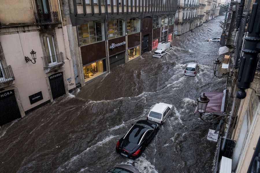 Floodwater flows down a street like a river, splashing around parked vehicles.
