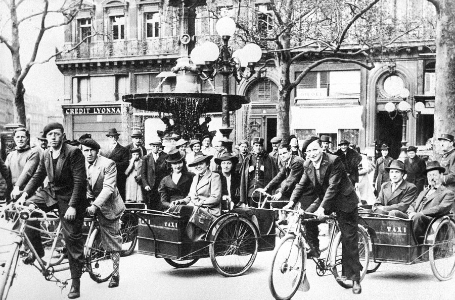 Onlookers watch as two pedal-taxis carry passengers in small carriages towed behind them.