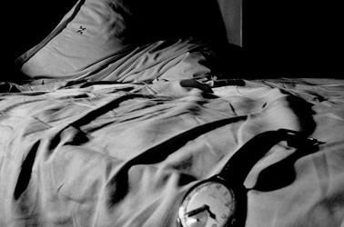 In a black-and-white photo, a watch hangs off an unmade bed cast in shadows.