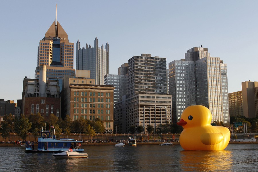 A huge inflatable rubber duck is towed up a river, past tall buildings.