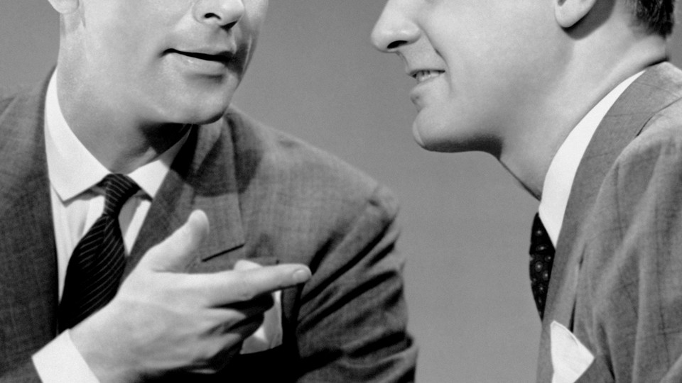 A black and white close up of two men in suits talking; one points at the other in a "you got it" sort of gesture