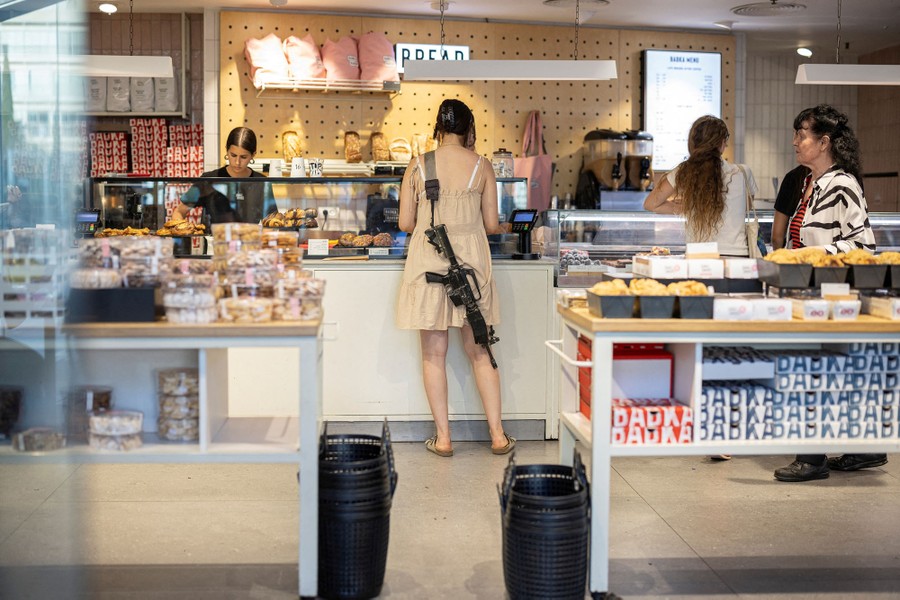 A woman in civilian clothing carries a rifle on a shoulder strap while shopping in a bakery.