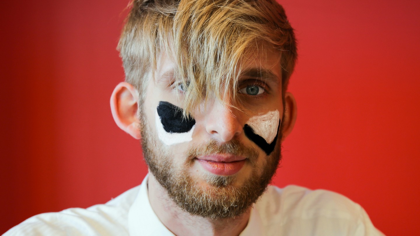 Robinson Meyer, a white man with blond hair and a beard, looking at the camera with white and black triangles painted on his cheeks. He is against a red background.