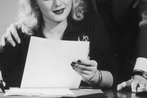 Black-and-white photo of a man putting his hand on a woman's shoulder and leaning over behind her
