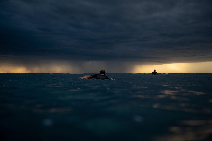 Two people paddle on surfboards with a thunderstorm and lightning bolt visible in the distance.