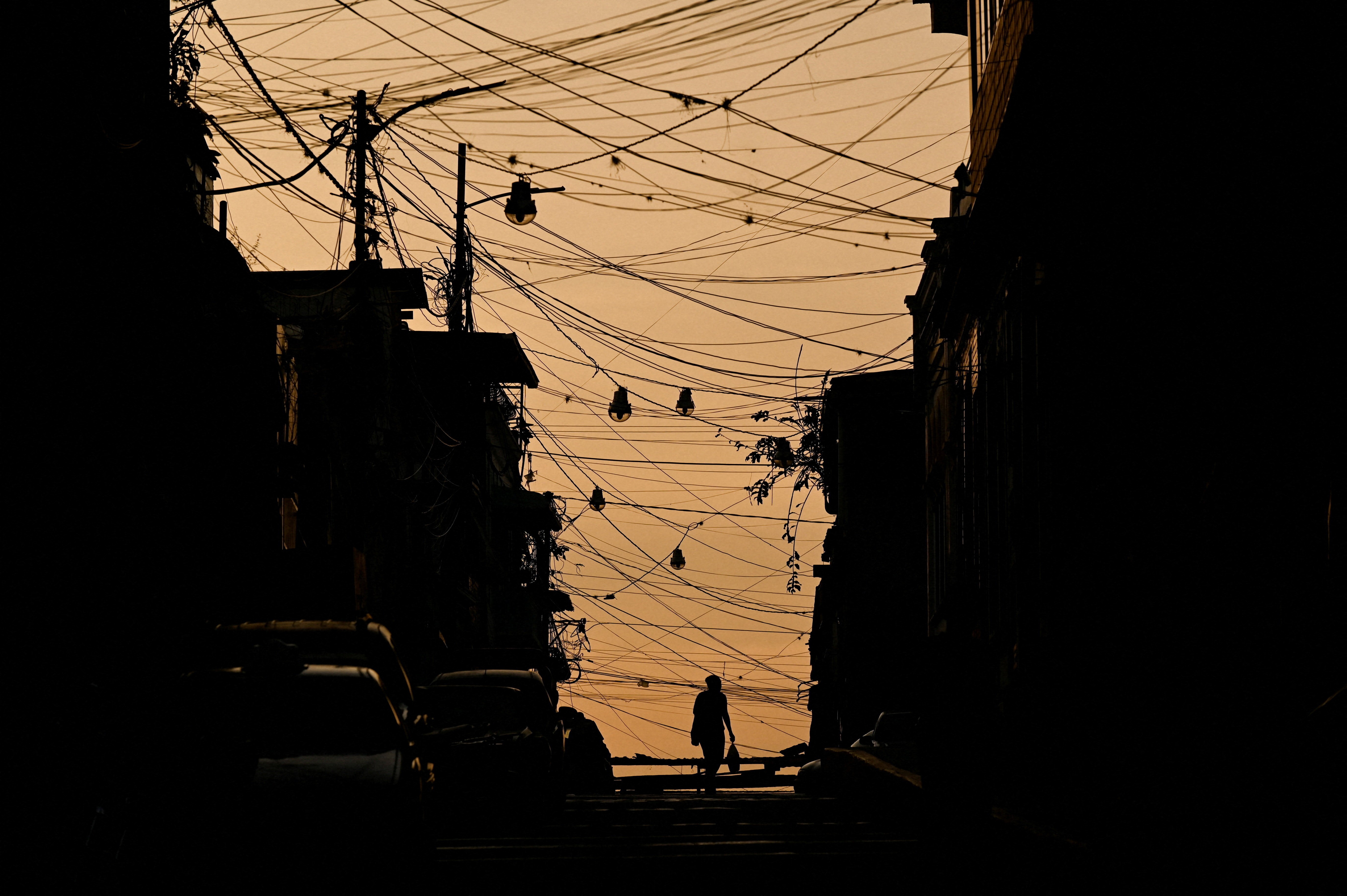 A woman walks with a shopping bag through a city street covered by a web of power lines, as the sun sets.