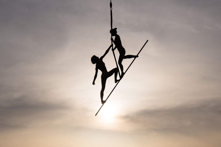 Two people perform midair on a thin bar suspended from a rope.