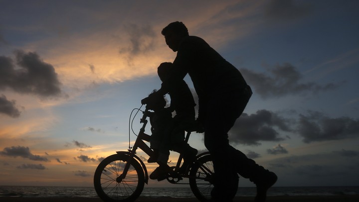 A man guides a child riding a two-wheel bike. It's sunset, and the figures appear as silhouettes.