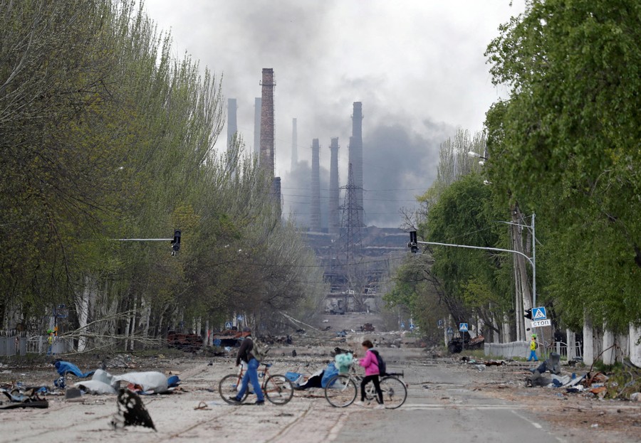 Two people walk their bicycles across a rubble-strewn street, with a factory and black smoke visible in the distance.