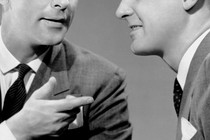 A black and white close up of two men in suits talking; one points at the other in a "you got it" sort of gesture