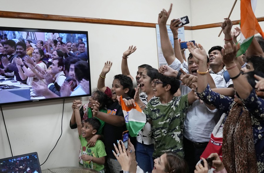 A crowd of children and adults cheer while looking at a television screen showing cheering team members of a successful space mission.