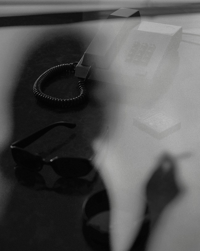 A black-and-white image of a woman's shadow over a desk. The desk has a phone and sunglasses on it.