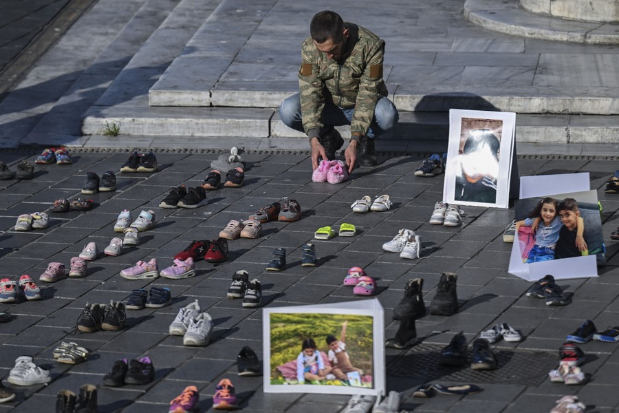 A person kneels down beside dozens of pairs of children's shoes placed in a city square as a protest.