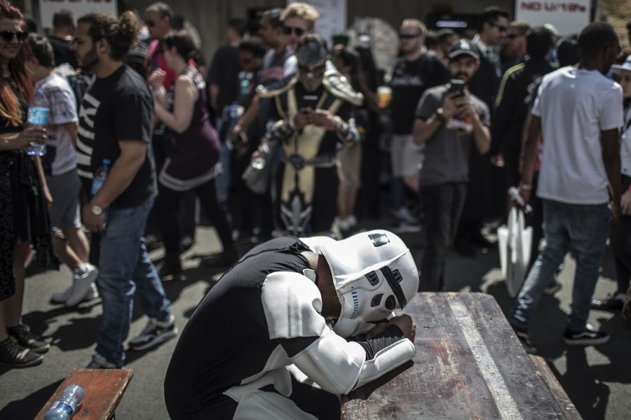 A cosplayer dressed as a stormtrooper naps at a picnic table near a crowd of people.