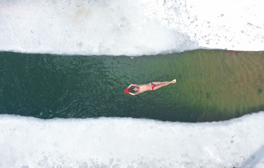 An aerial view of a winter swimming enthusiast floating in a partly-frozen lake.