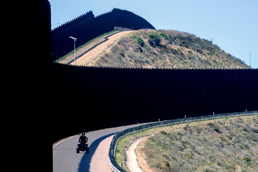 A border-patrol agent rides an ATV along a section of tall border fence.