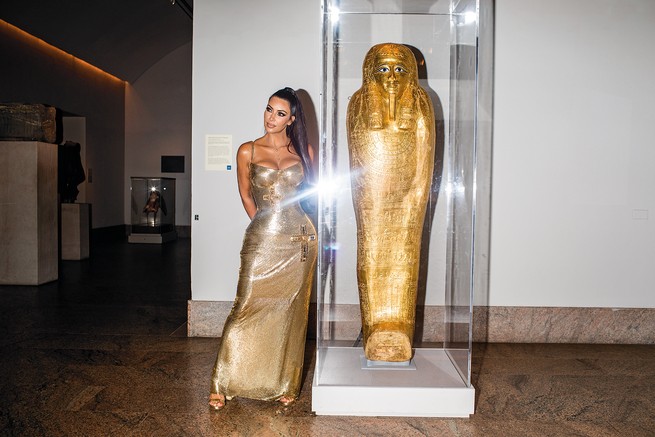 photo of Kim Kardashian in gold evening gown standing next to glass case with golden Egyptian coffin
