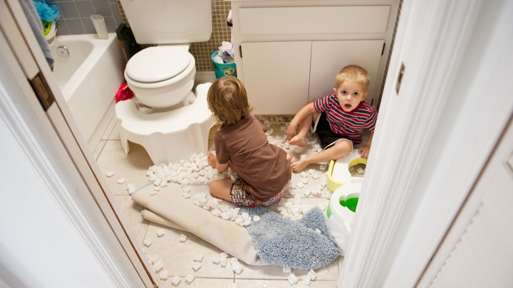 Two young children sit on the floor of a bathroom surrounded by packing peanuts. One child looks clearly alarmed and perhaps guilty of misbehavior.