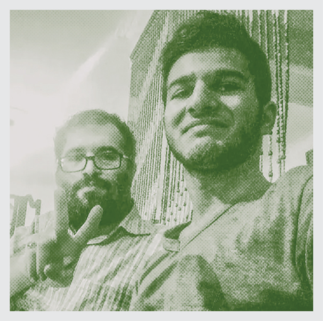 عکس بی‌تکلف مردی ریشو با عینک در پس‌زمینه که دو انگشت خود را بالا گرفته و مردی جوان‌تر در پیش‌زمینه لبخند می‌زند