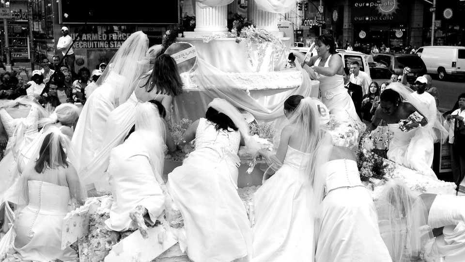 A black-and-white image of brides in wedding dresses scrambling onto a large structural wedding display