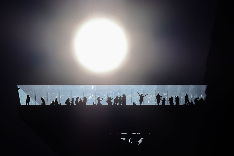 The silhouettes of a couple dozen people are seen on a high observation deck, with the sun in the background.