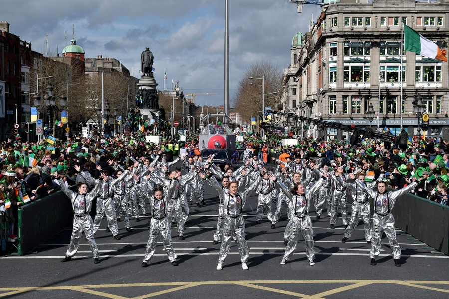 A group of performers wearing silver costumes stand with arms raised in a parade.