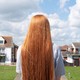 A white girl with long red hair, wearing a gray t-shirt, has her back to the camera and looks out at two lawns and two houses on a suburban street.
