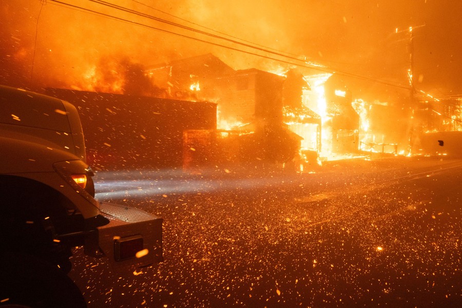 Embers flying across a road as houses burn in the background.