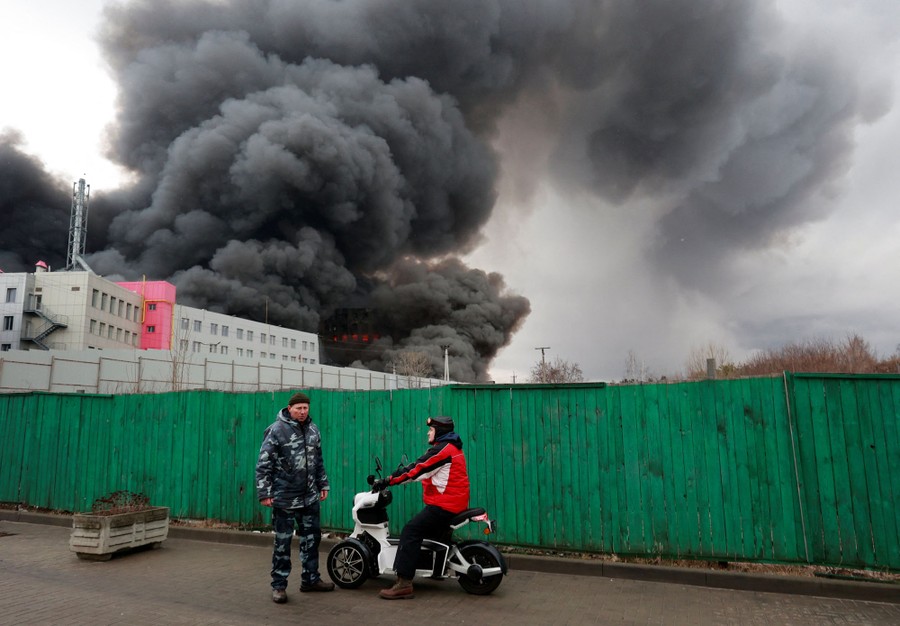 A person on a scooter talks with another person on a street alongside a fence. Behind them a large building burns, sending black smoke into the sky.